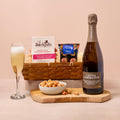 Bottle of L'Extra Langlois champagne, glass of champagne, bowl of popcorn, and basket of snacks on a wooden board with a pink background.