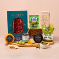 Assorted food products including a box of Tempus salami, Olly's pickles, and other snacks on a wooden board with a beige background.