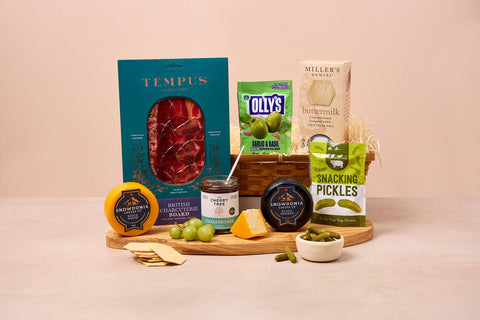 Assorted food products including a box of Tempus salami, Olly's pickles, and other snacks on a wooden board with a beige background.
